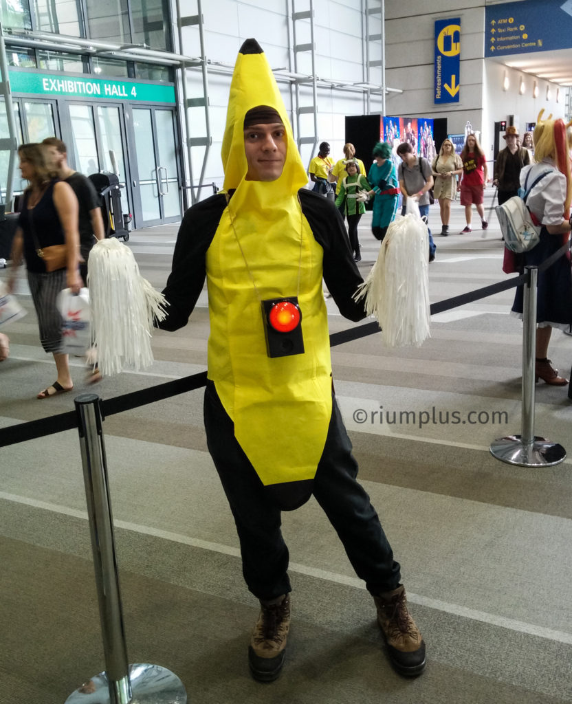 A man wearing a bright yellow banana costume, with white pom-poms in each hand. A black box with a big red button & speaker is dangling around his neck. He's doing a Peanut Butter Jelly Time cosplay