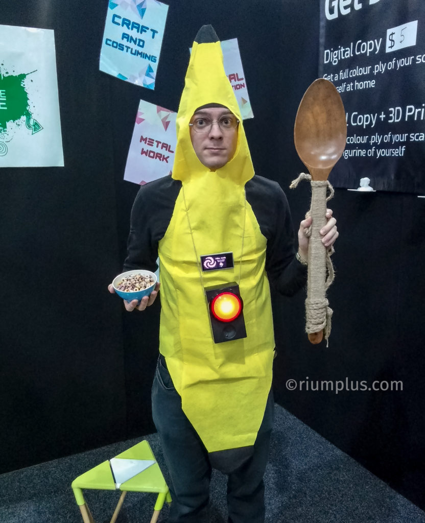 A man wearing a bright yellow banana costume, carrying a small bowl of cereal in one hand & a 75cm long wooden spoon in the other. A black box with a big red button & speaker is dangling around his neck.