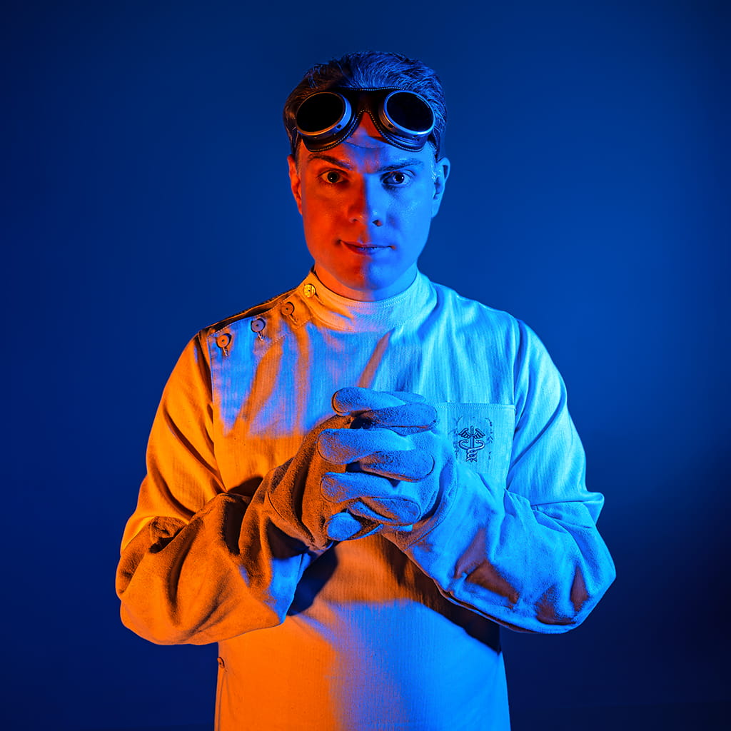 Mike Ando aka riumplus aka RIUM+, a man with short brown hair. He's wearing a vintage medical smock, thick white leather gloves, and goggles on his forehead. The whole scene is lit orange and blue like is common in current scifi movie posters.