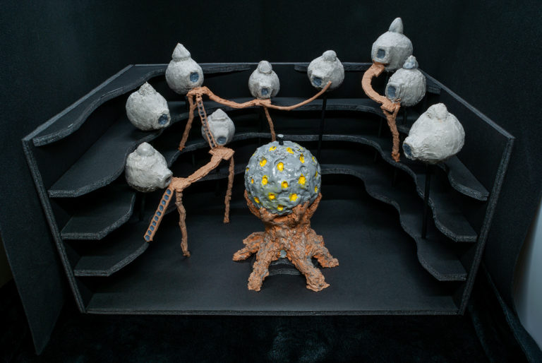 A decorative set of cake pops in a custom black stand, inspired by the video game Riven: The Sequel to Myst. The main one in the middle is Tay, which looks vaguely like a golf ball on a tree trunk. Around the “cliff” edges are little villager hut cake pops. These have miniature chocolate bridges & ladders between them